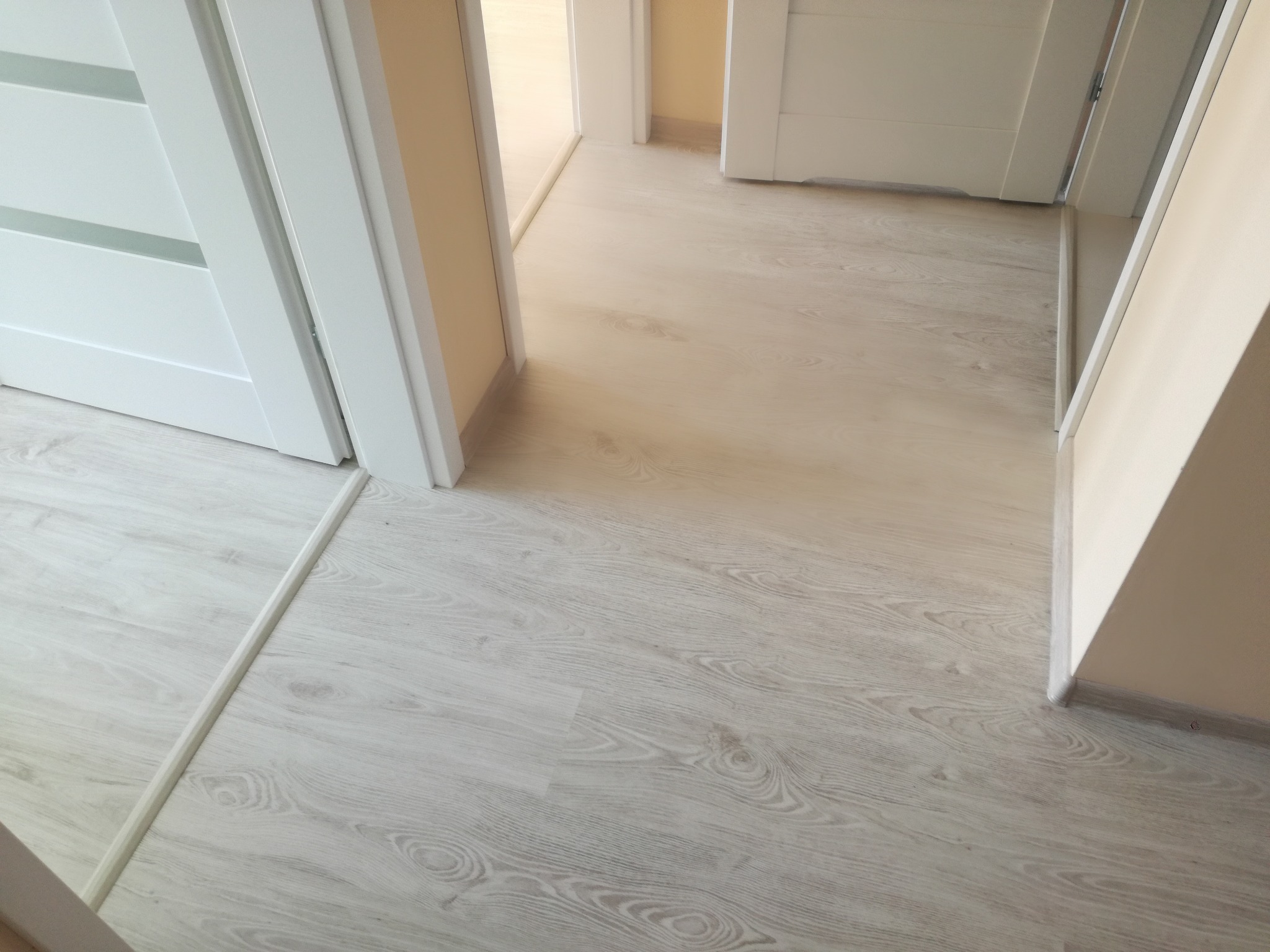 Jasne panele podłogowe z widocznym wzorem drewna, białe drzwi w tle, widok na korytarz.