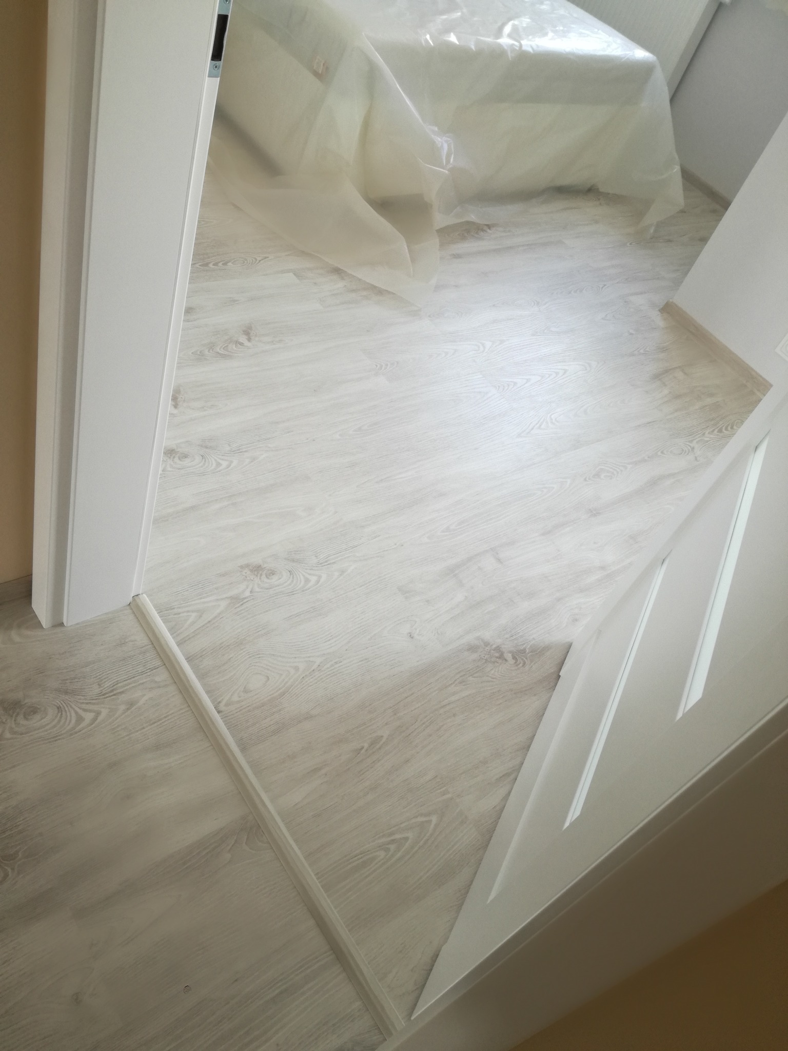 Jasne panele podłogowe ułożone w pokoju, widoczne zza otwartych białych drzwi, z łóżkiem przykrytym folią ochronną w tle.