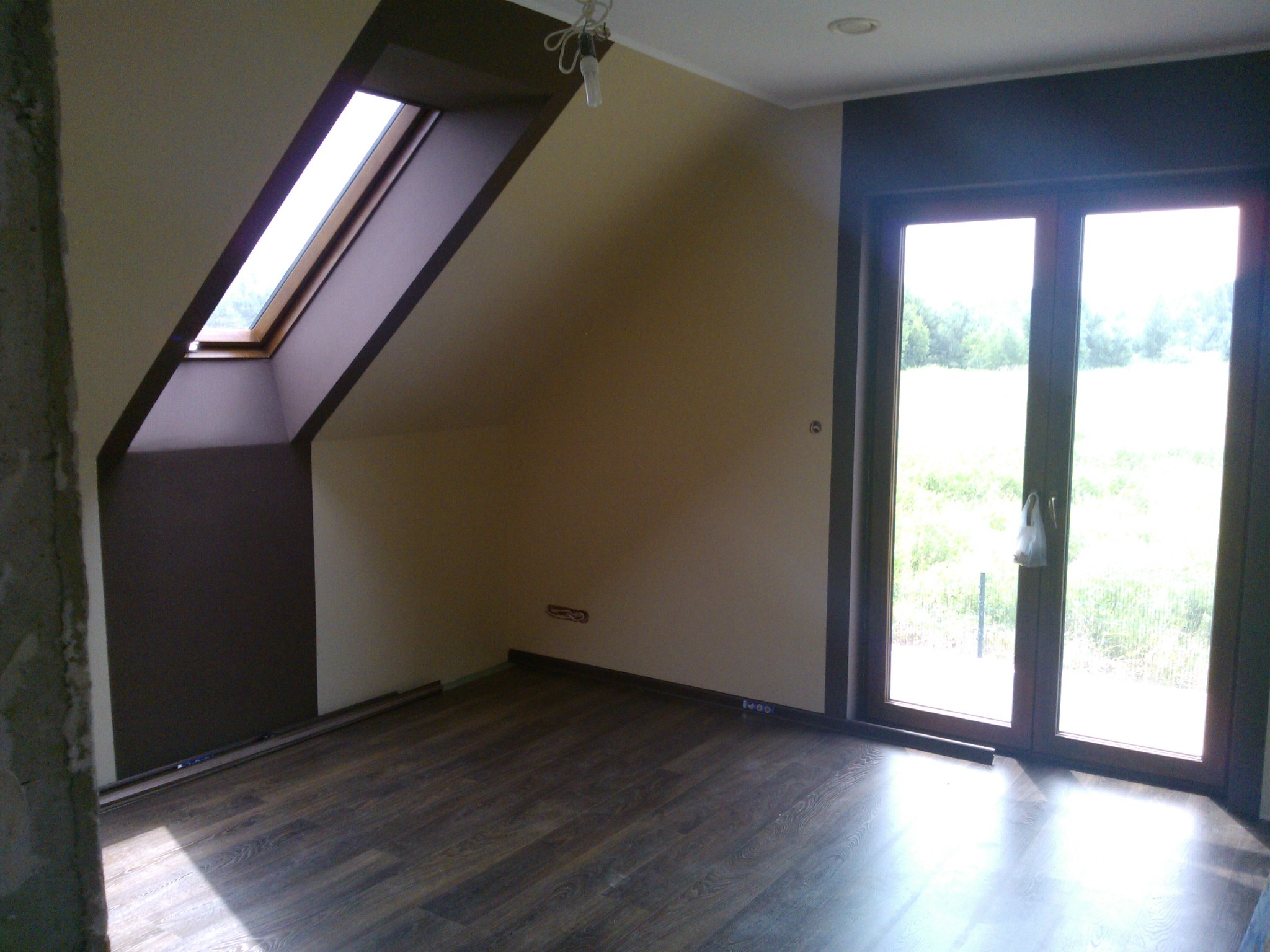 Pokój na poddaszu z ułożoną podłogą z paneli, widoczne okno dachowe, drzwi balkonowe i ściany pomalowane na dwa kolory. Fragment ściany w stanie surowym.