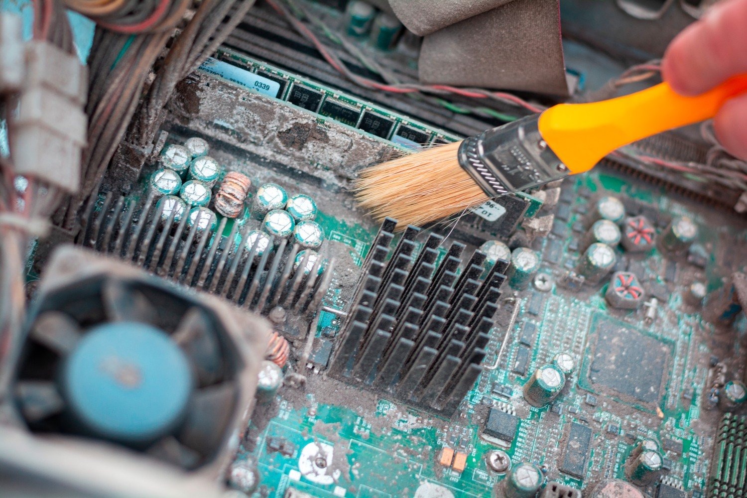 Szczoteczka z pomarańczową rączką czyści zakurzoną płytę główną komputera, widoczne kondensatory, radiator i gniazda pamięci RAM.