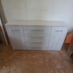 Komoda z jasnego drewna z czterema szufladami i dwojgiem drzwi, z metalowymi uchwytami, ustawiona na podłodze w surowym wnętrzu.