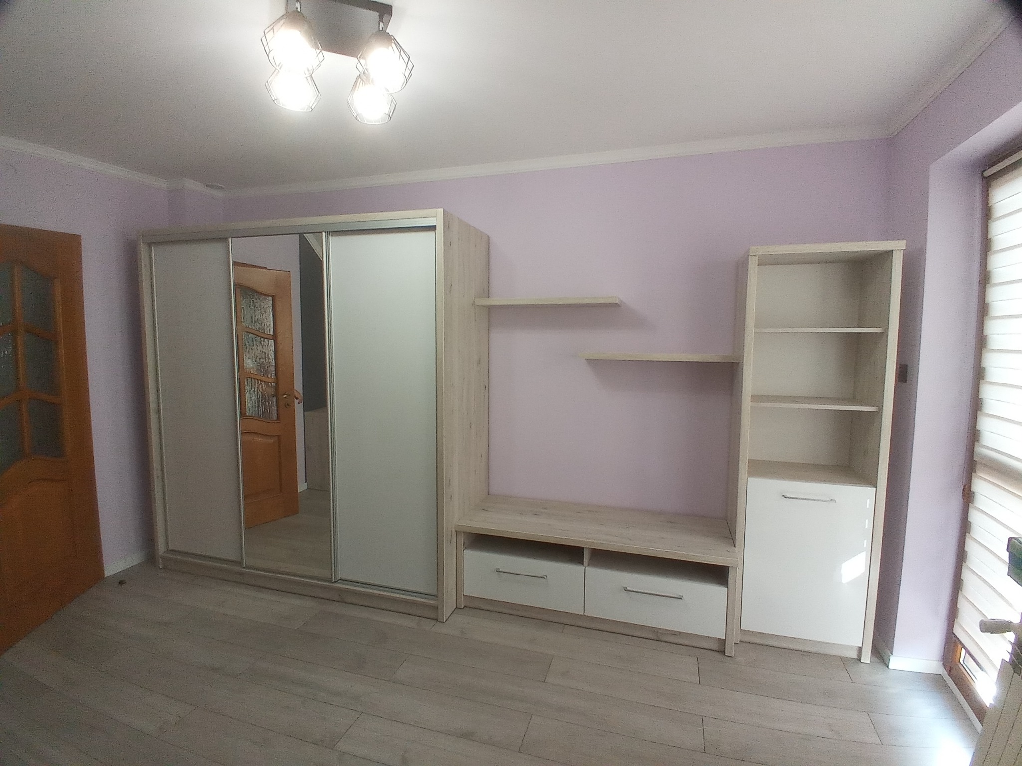 Jasna szafa przesuwna z lustrem i regał z półkami w pokoju z fioletowymi ścianami i drewnianą podłogą.