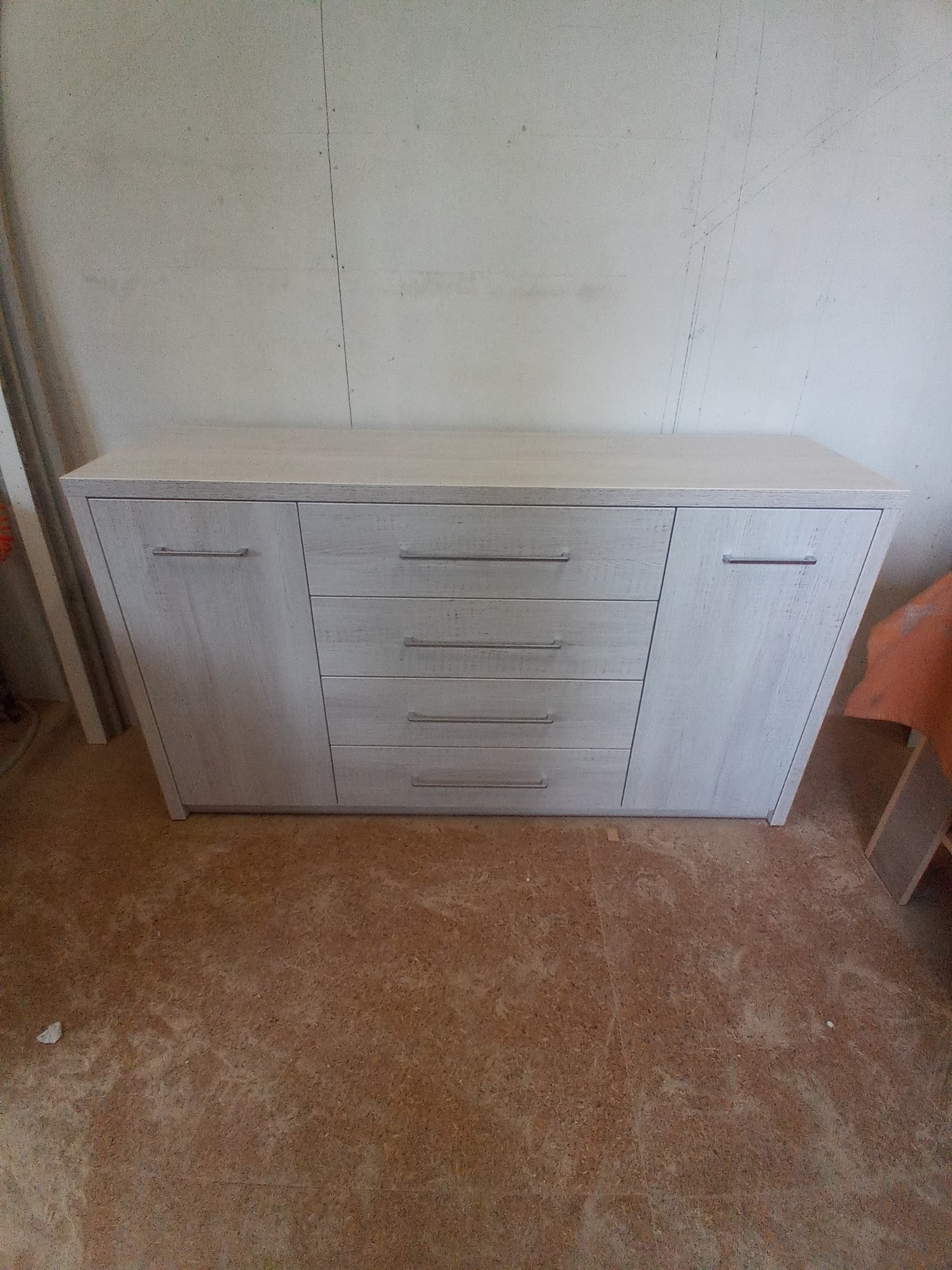 Komoda z jasnego drewna z czterema szufladami i dwojgiem drzwi, z metalowymi uchwytami, ustawiona na podłodze w surowym wnętrzu.