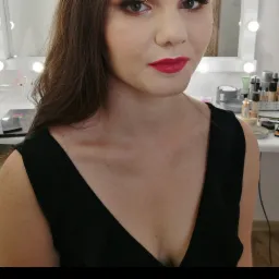 Portret kobiety z intensywnym makijażem oczu i jaskrawoczerwoną szminką, wykonany w salonie kosmetycznym z lustrem podświetlanym żarówkami w tle.