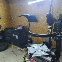 Wnętrze warsztatu z maszyną do wyważania kół Grubber EM760 i montażownicą do opon Wulcan CE, stos opon przy ścianie z płyty OSB, oświetlenie jarzeniowe.