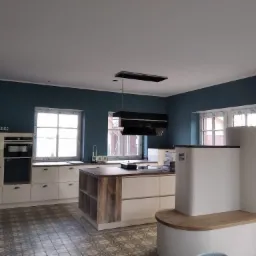 Wnętrze kuchni po remoncie z wyspą, kremowe szafki, ściany w kolorze petrolu, dekoracyjna podłoga z płytek, nowoczesny okap i kominek kaflowy z drewnianym blatem.