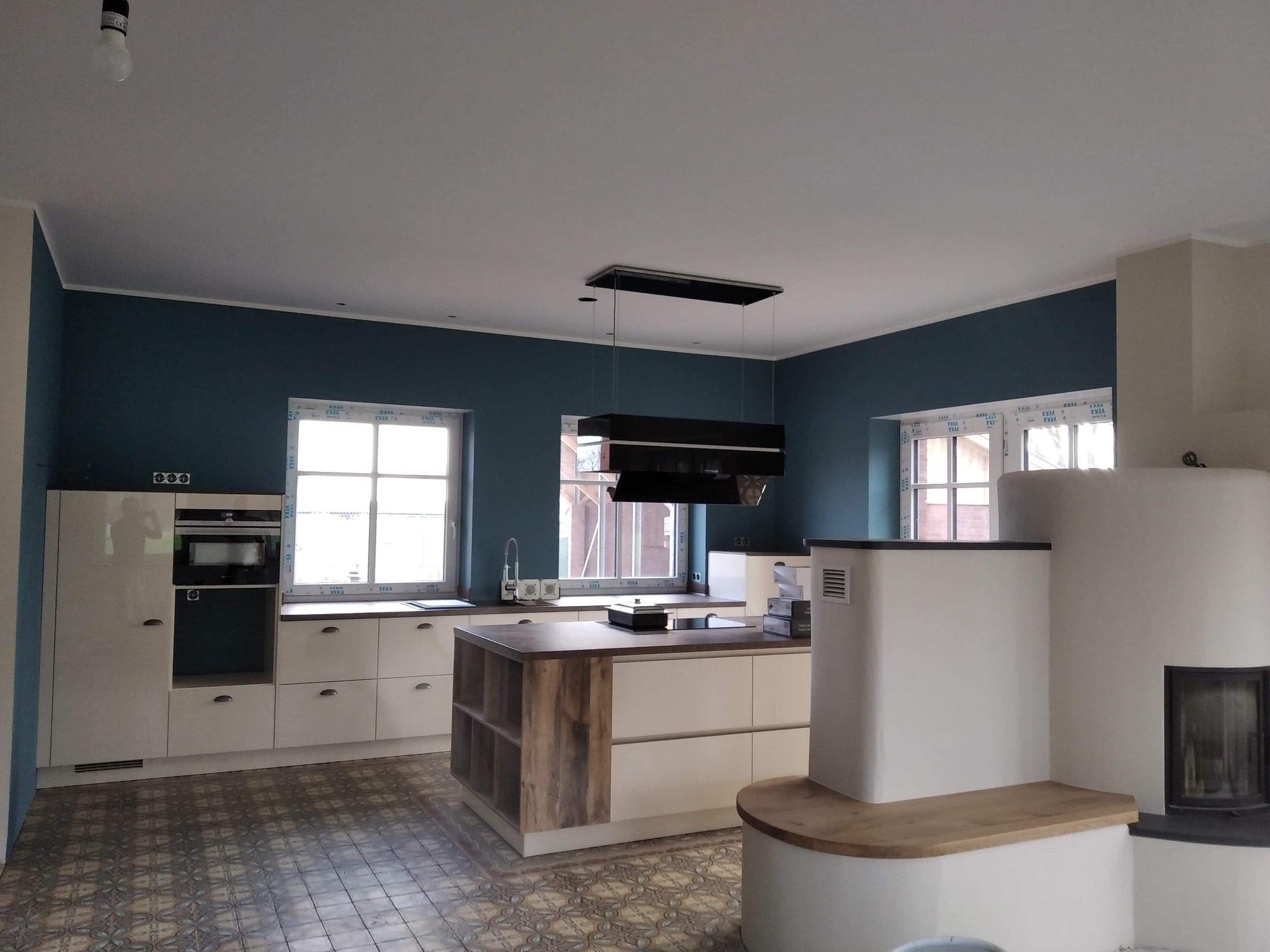 Wnętrze kuchni po remoncie z wyspą, kremowe szafki, ściany w kolorze petrolu, dekoracyjna podłoga z płytek, nowoczesny okap i kominek kaflowy z drewnianym blatem.