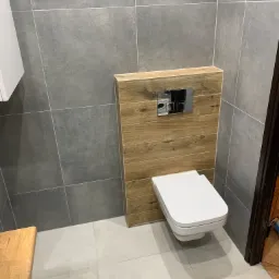 Nowoczesna toaleta podwieszana z drewnianą obudową stelaża, chromowany przycisk spłukujący, szare płytki ścienne i podłogowe.