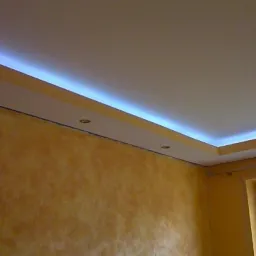 Nowoczesny sufit podwieszany z oświetleniem LED wzdłuż krawędzi i wbudowanymi punktowymi lampami, ściany w odcieniach żółci i pomarańczy, widoczne okno z ramą PCV.