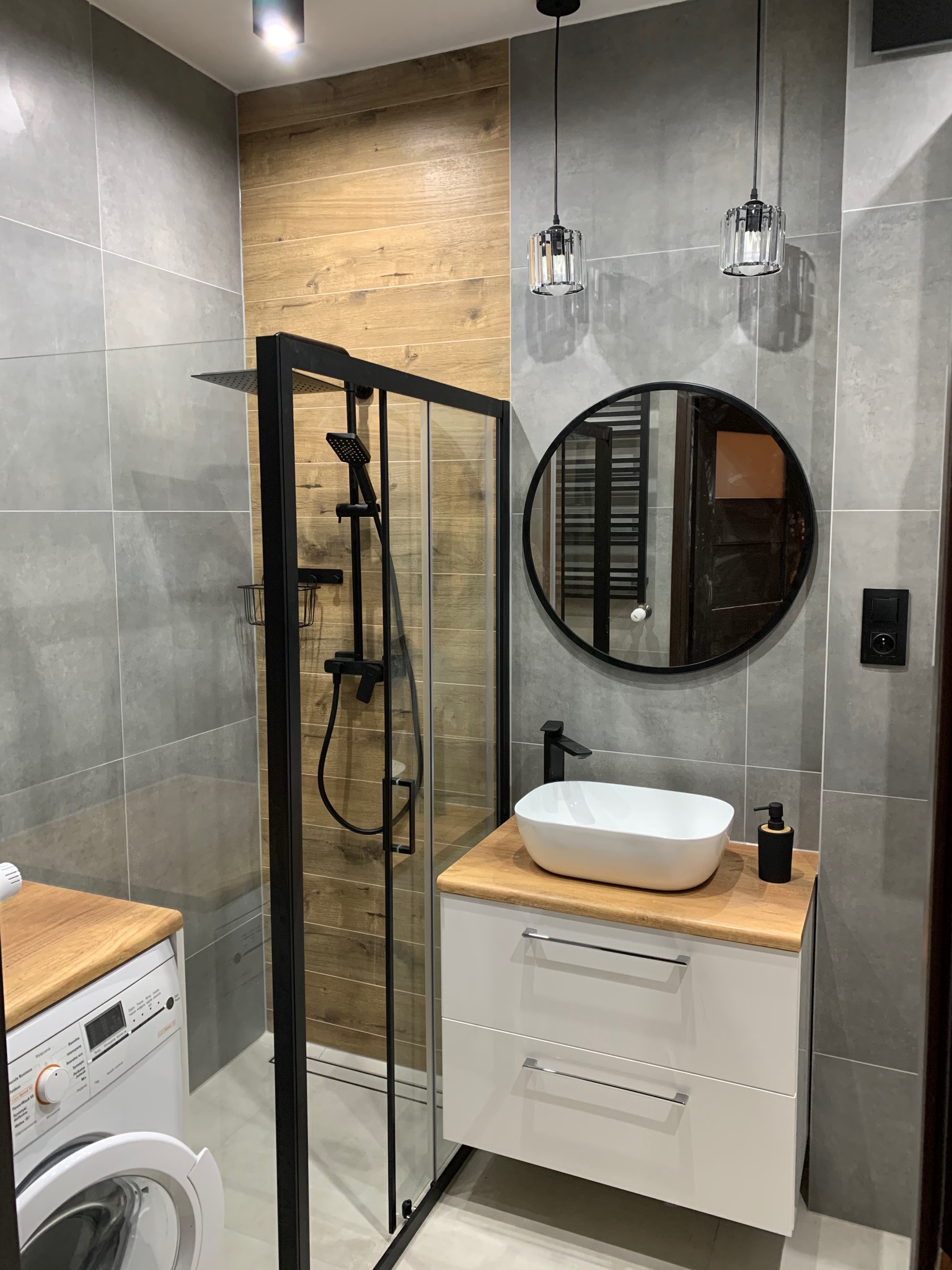Nowoczesna łazienka z szarą płytką, drewnopodobną ścianą, kabiną prysznicową z czarnymi profilami, okrągłym lustrem i umywalką nablatową na szafce z drewnianym blatem, oświetlona wiszącymi lampami.