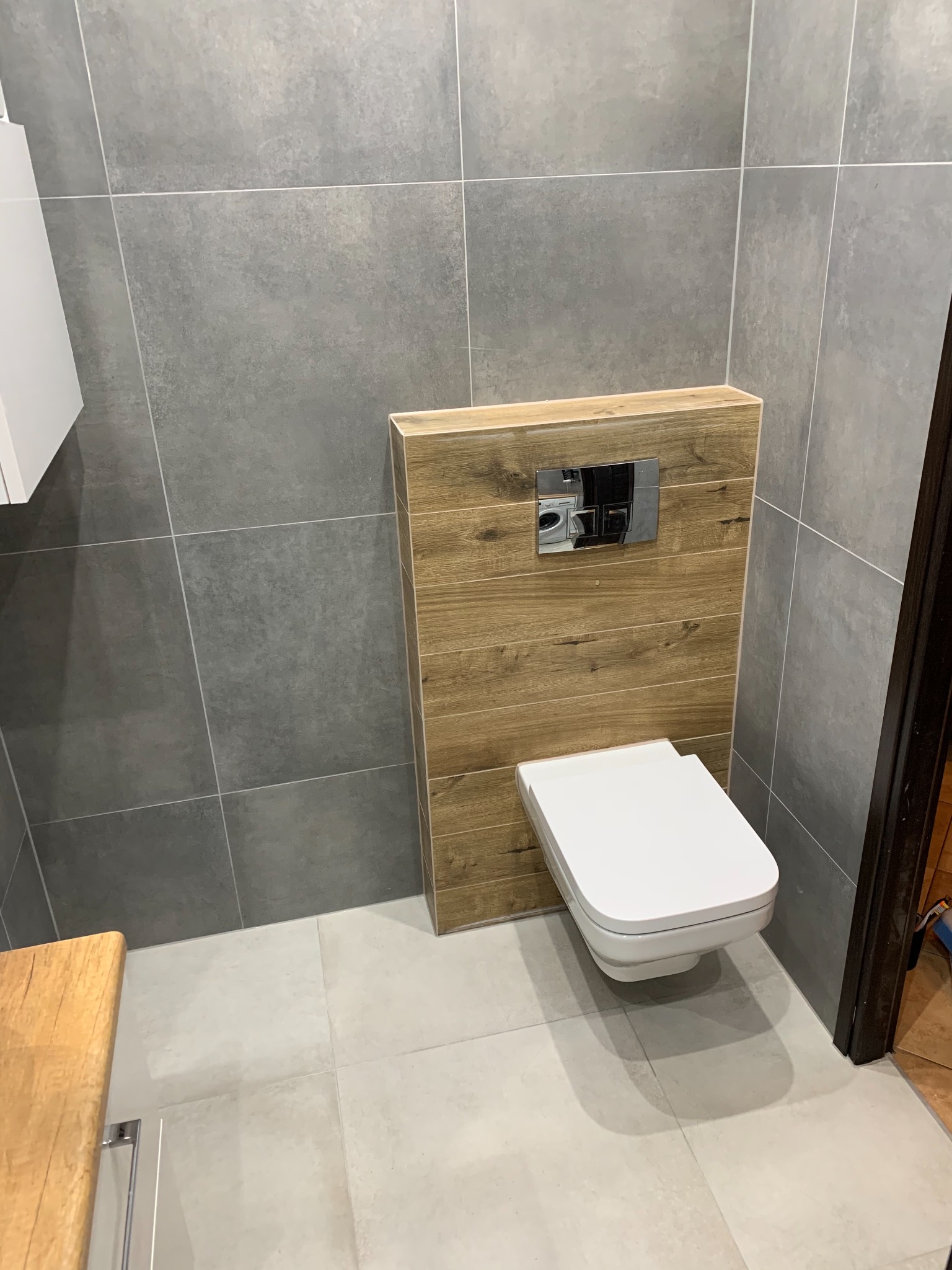Nowoczesna toaleta podwieszana z drewnianą obudową stelaża, chromowany przycisk spłukujący, szare płytki ścienne i podłogowe.