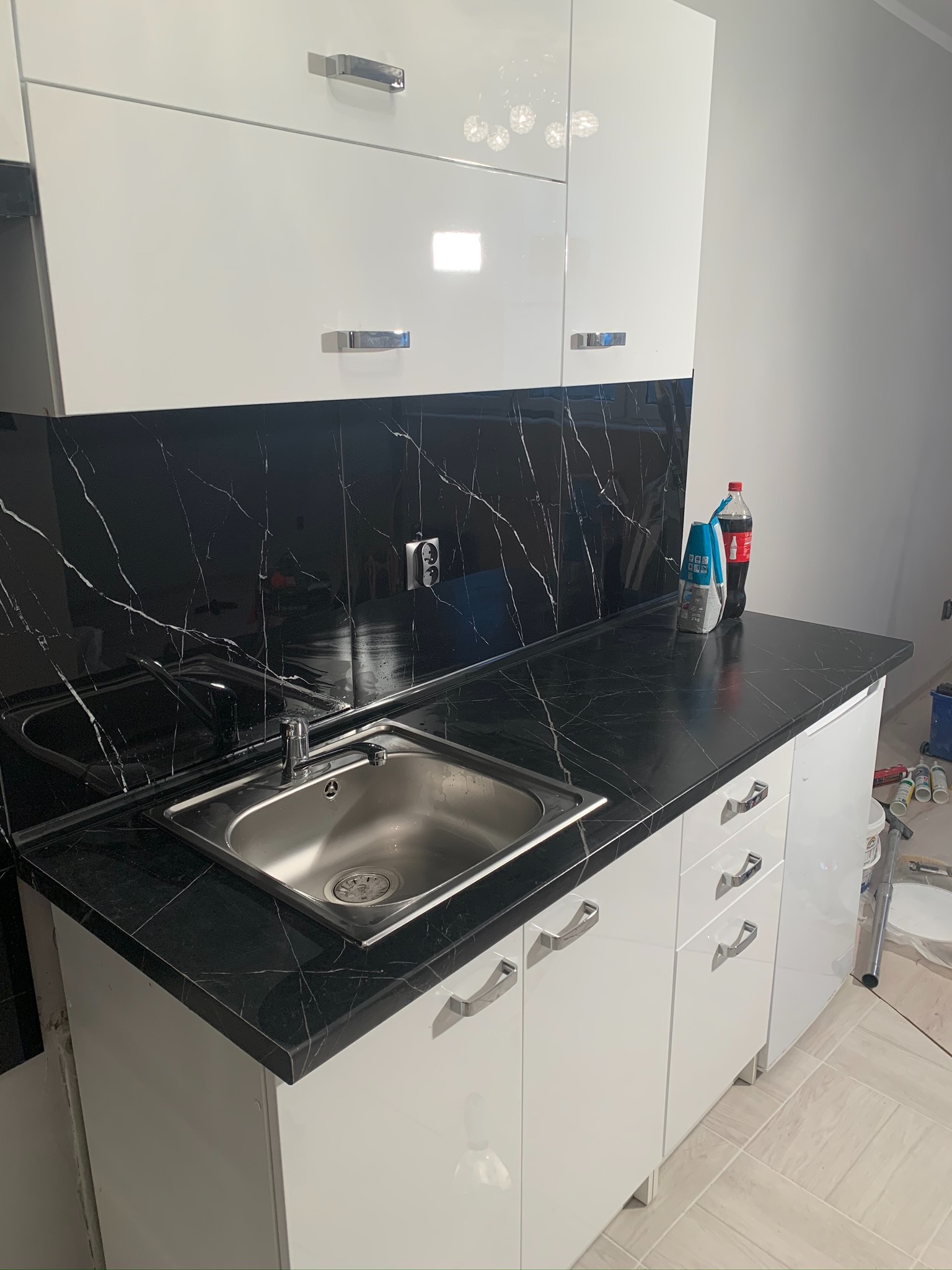 Nowoczesna kuchnia z białymi szafkami i czarnym blatem imitującym marmur, zlewozmywak stalowy, butelka Coca-Coli i worek z klejem na blacie, widoczne narzędzia malarskie w tle.