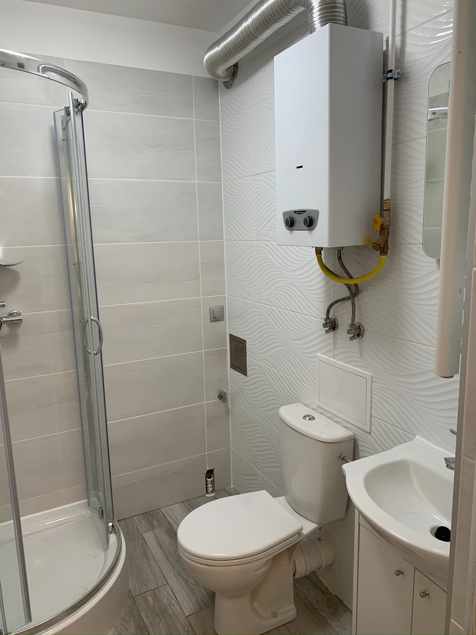 Wykończona łazienka z kabiną prysznicową, toaletą, umywalką z szafką i zamontowanym piecykiem gazowym Ariston na ścianie z dekoracyjnymi płytkami.