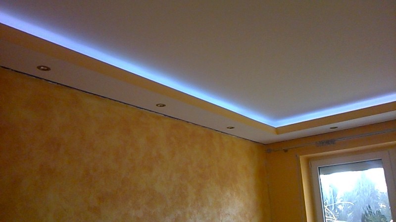 Nowoczesny sufit podwieszany z oświetleniem LED wzdłuż krawędzi i wbudowanymi punktowymi lampami, ściany w odcieniach żółci i pomarańczy, widoczne okno z ramą PCV.