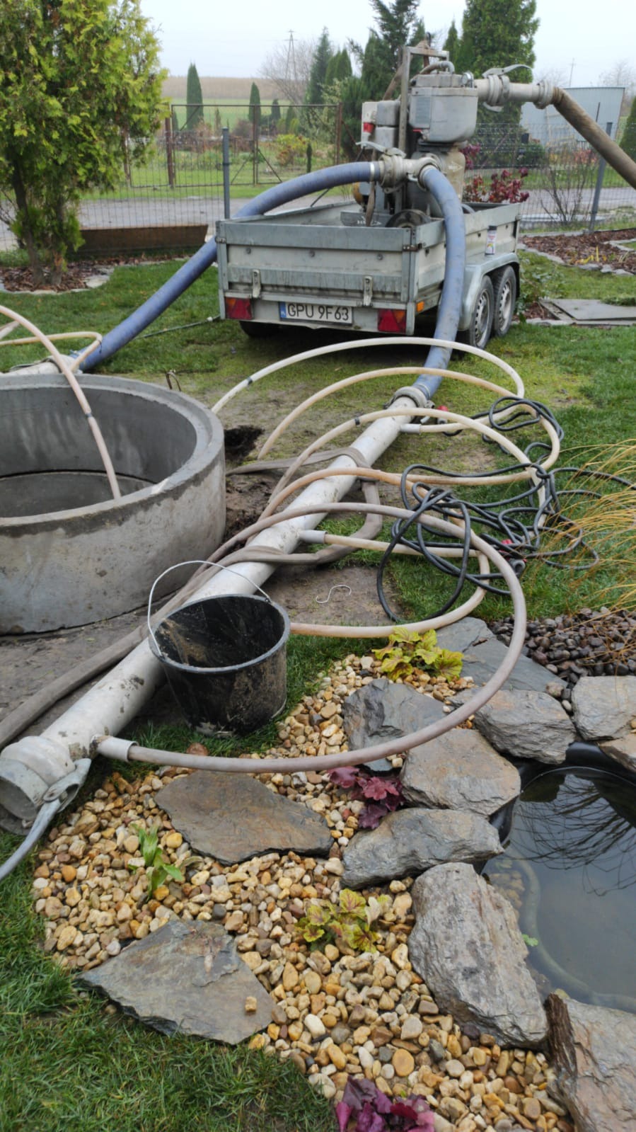 Instalacja odwadniająca metodą igłofiltrową w ogrodzie: widoczny betonowy krąg studni, pompa na przyczepie z niebieskimi wężami, oraz oczko wodne z kamieniami i roślinami.