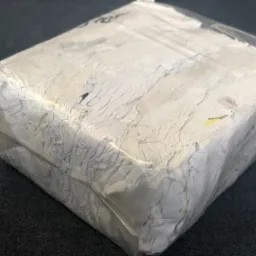 Zwarta kostka białej, pogniecionej tkaniny, prawdopodobnie bawełny, zapakowana w przezroczystą folię, leżąca na ciemnoszarym tle.