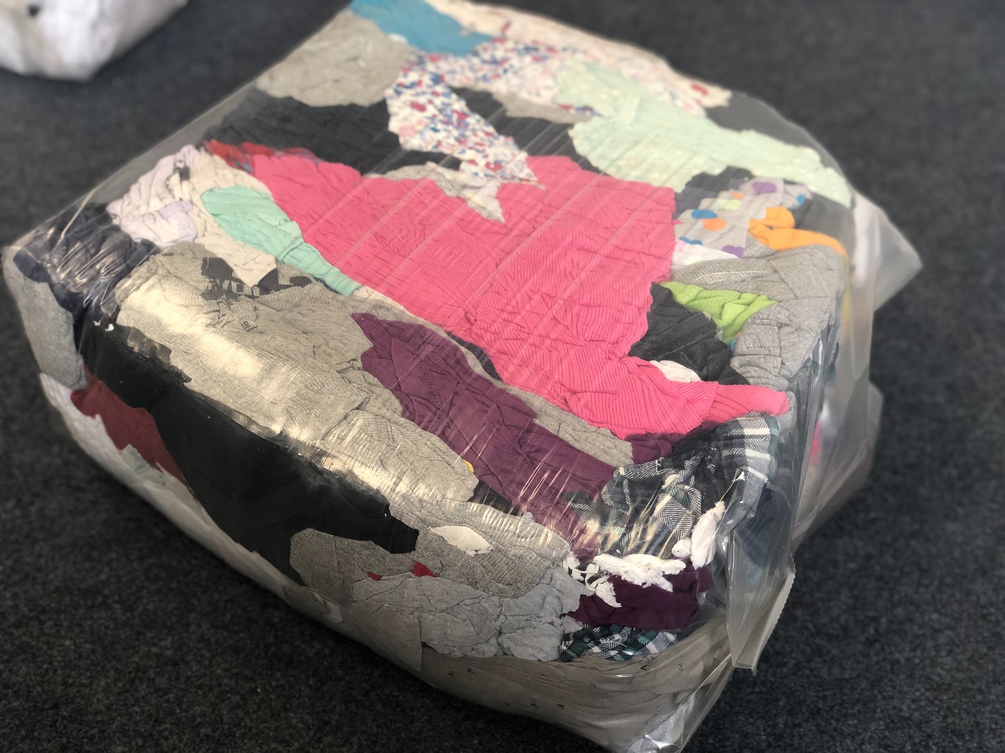 Duży pakiet z różnorodną, kolorową odzieżą zapakowaną w przezroczystą folię, leżący na szarym dywanie.