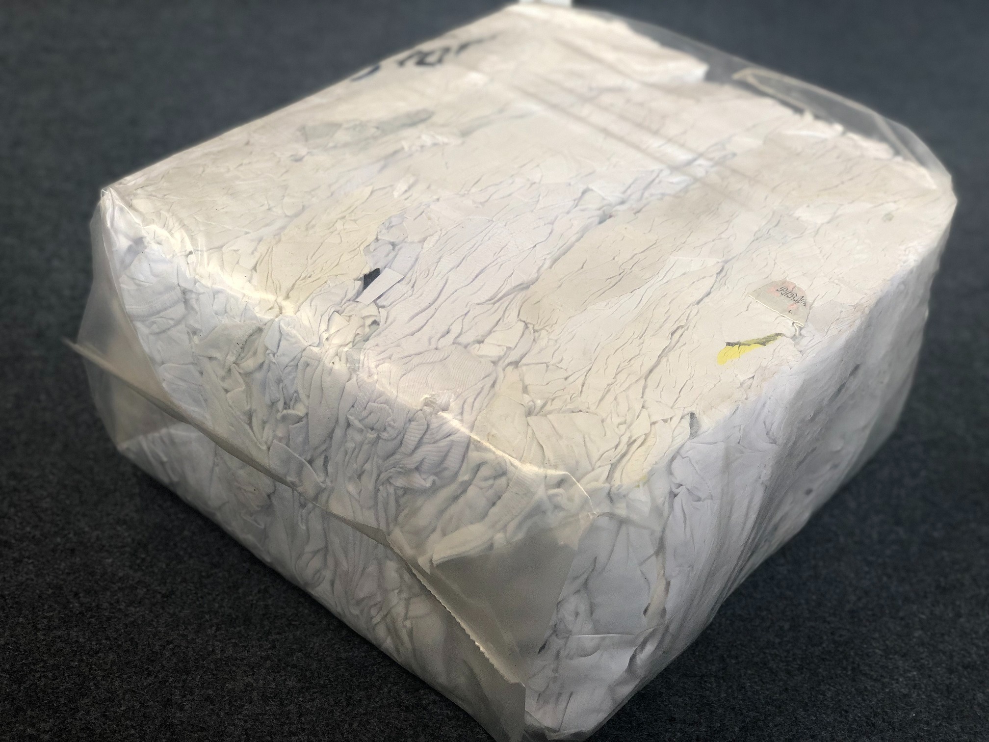 Zwarta kostka białej, pogniecionej tkaniny, prawdopodobnie bawełny, zapakowana w przezroczystą folię, leżąca na ciemnoszarym tle.