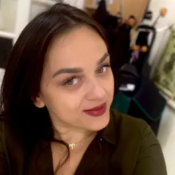 Portret kobiety z ciemnymi włosami i zielonymi oczami, w zielonej koszuli, z delikatnym makijażem i złotym naszyjnikiem z symbolem Barana, spoglądającej w obiektyw.