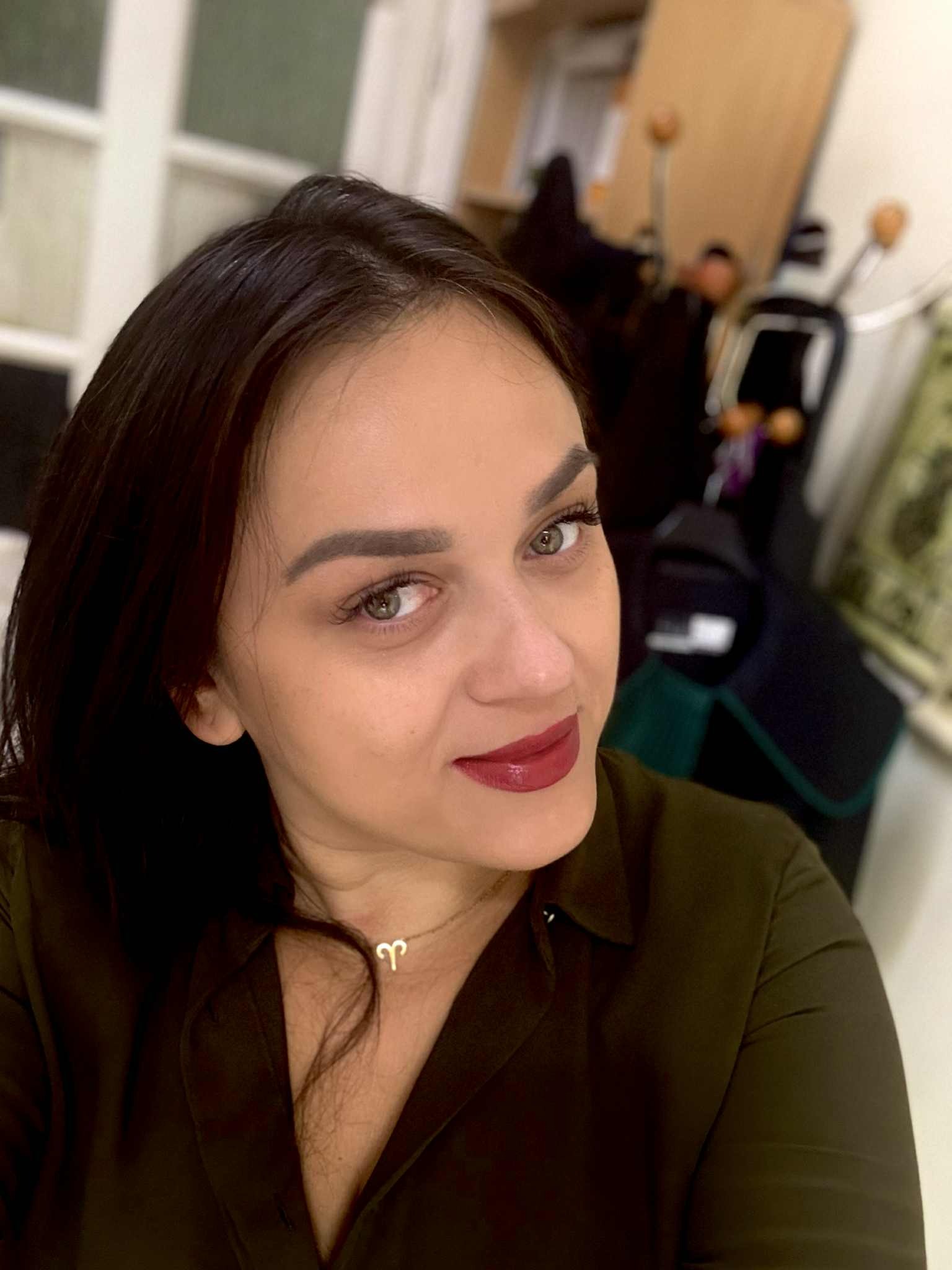 Portret kobiety z ciemnymi włosami i zielonymi oczami, w zielonej koszuli, z delikatnym makijażem i złotym naszyjnikiem z symbolem Barana, spoglądającej w obiektyw.