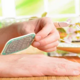 Elektrostymulacja: osoba aplikuje elektrodę na przedramię, widoczny kabel urządzenia na tle okna i kwiatów.