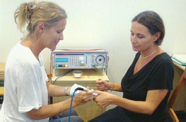 Terapeutka wykonuje zabieg biorezonansu na dłoni pacjentki przy użyciu specjalistycznego urządzenia z wyświetlaczem i elektrodami.