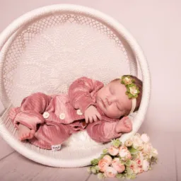 Śpiące nowonarodzone dziecko w różowym welurowym ubranku z dekoracją kwiatową na głowie, ułożone w okrągłym, plecionym koszu z koronkowym wykończeniem, obok bukiet bladoróżowych róż.