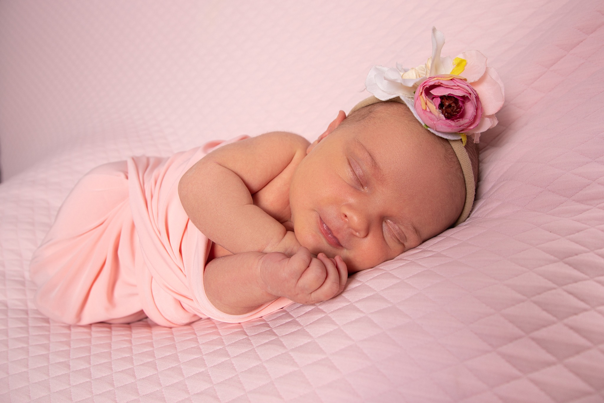 Śpiąca noworodka zawinięta w różowy materiał, z opaską z kwiatami na głowie, leżąca na różowej pikowanej poduszce.