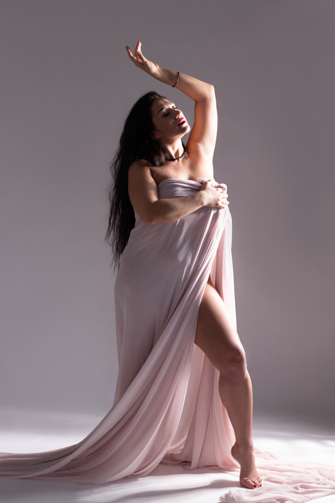 Kobieta w jasnoróżowej tkaninie, z uniesioną ręką, czerwone usta, czarne włosy, subtelne światło i cień, artystyczna poza.