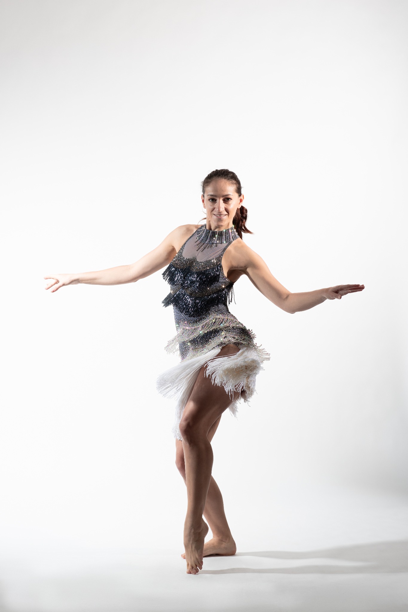Kobieta w błyszczącej sukience tanecznej z frędzlami, z rozłożonymi ramionami, na białym tle, w trakcie występu.