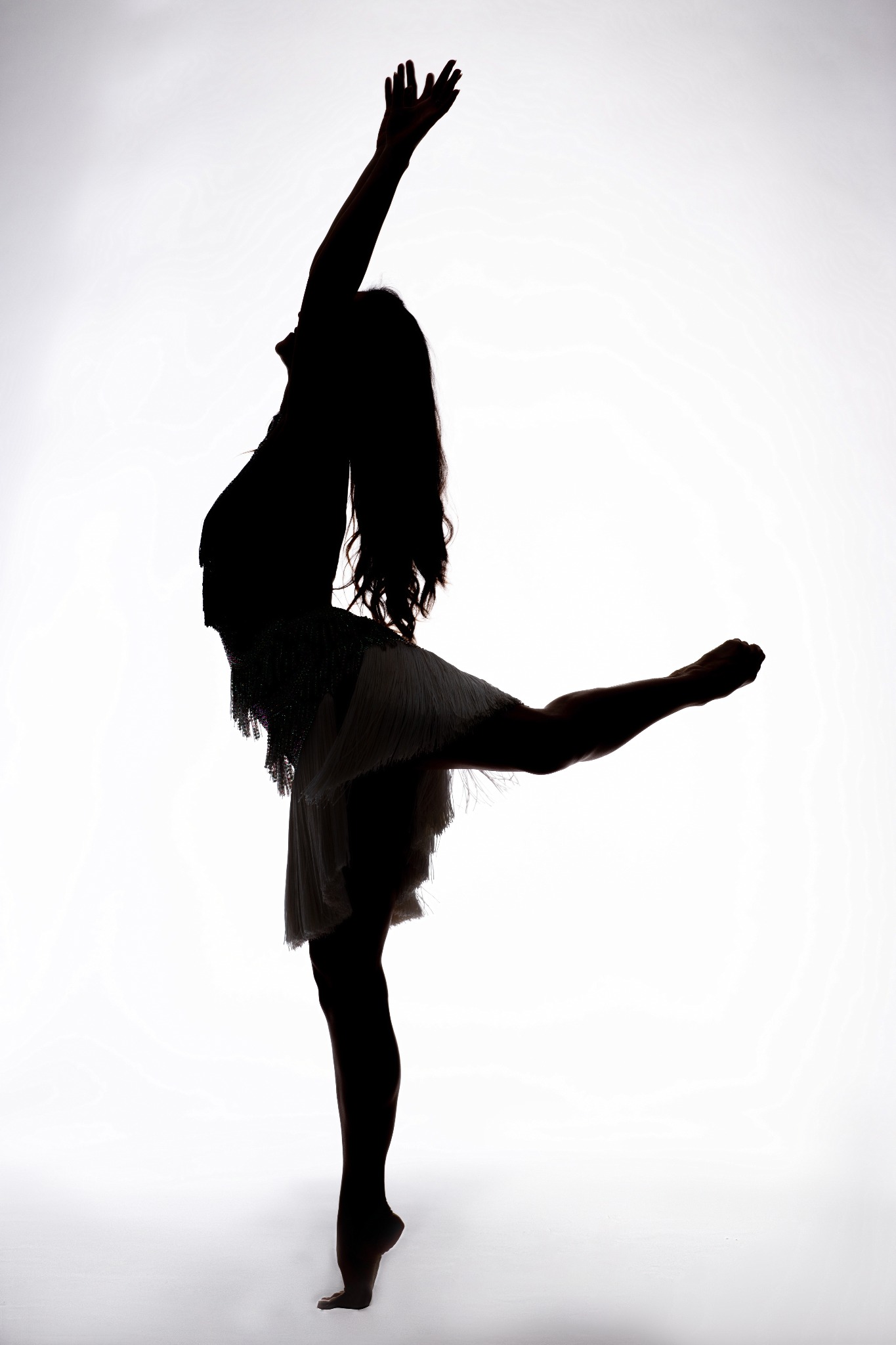 Sylwetka kobiety w tanecznej pozie na jasnym tle, z uniesioną ręką i wyciągniętą nogą, ubrana w strój z frędzlami.