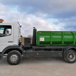 Ciężarówka Mercedes-Benz 1218 z zielonym kontenerem na platformie, widok z boku na tle pochmurnego nieba i krajobrazu.