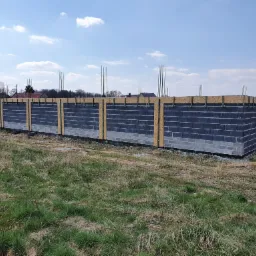 Wznoszenie ścian fundamentowych z bloczków betonowych, z widocznym deskowaniem i wystającymi prętami zbrojeniowymi, na tle łąki i błękitnego nieba.