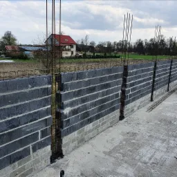 Wznoszenie ściany z szarych bloczków betonowych z pionowymi prętami zbrojeniowymi, widok na fundament i pobliskie budynki w tle.