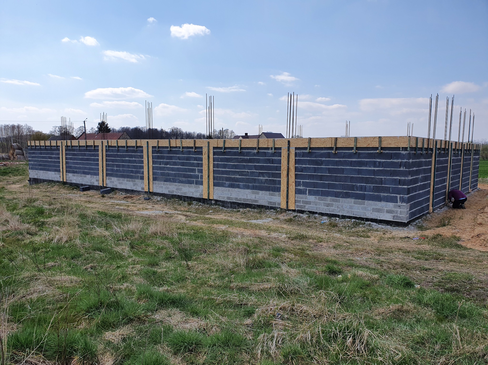 Wznoszenie ścian fundamentowych z bloczków betonowych, z widocznym deskowaniem i wystającymi prętami zbrojeniowymi, na tle łąki i błękitnego nieba.