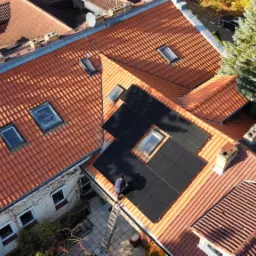 Montaż paneli słonecznych na dachu z czerwonej dachówki, widok z góry na instalatora pracującego obok okna dachowego, drabina oparta o dach, drzewo w tle.