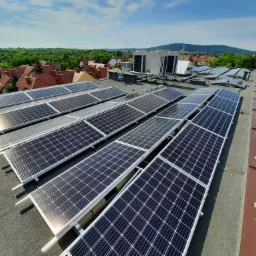 Rząd paneli fotowoltaicznych na płaskim dachu budynku, widok z góry, w tle drzewa, zabudowania i błękitne niebo.