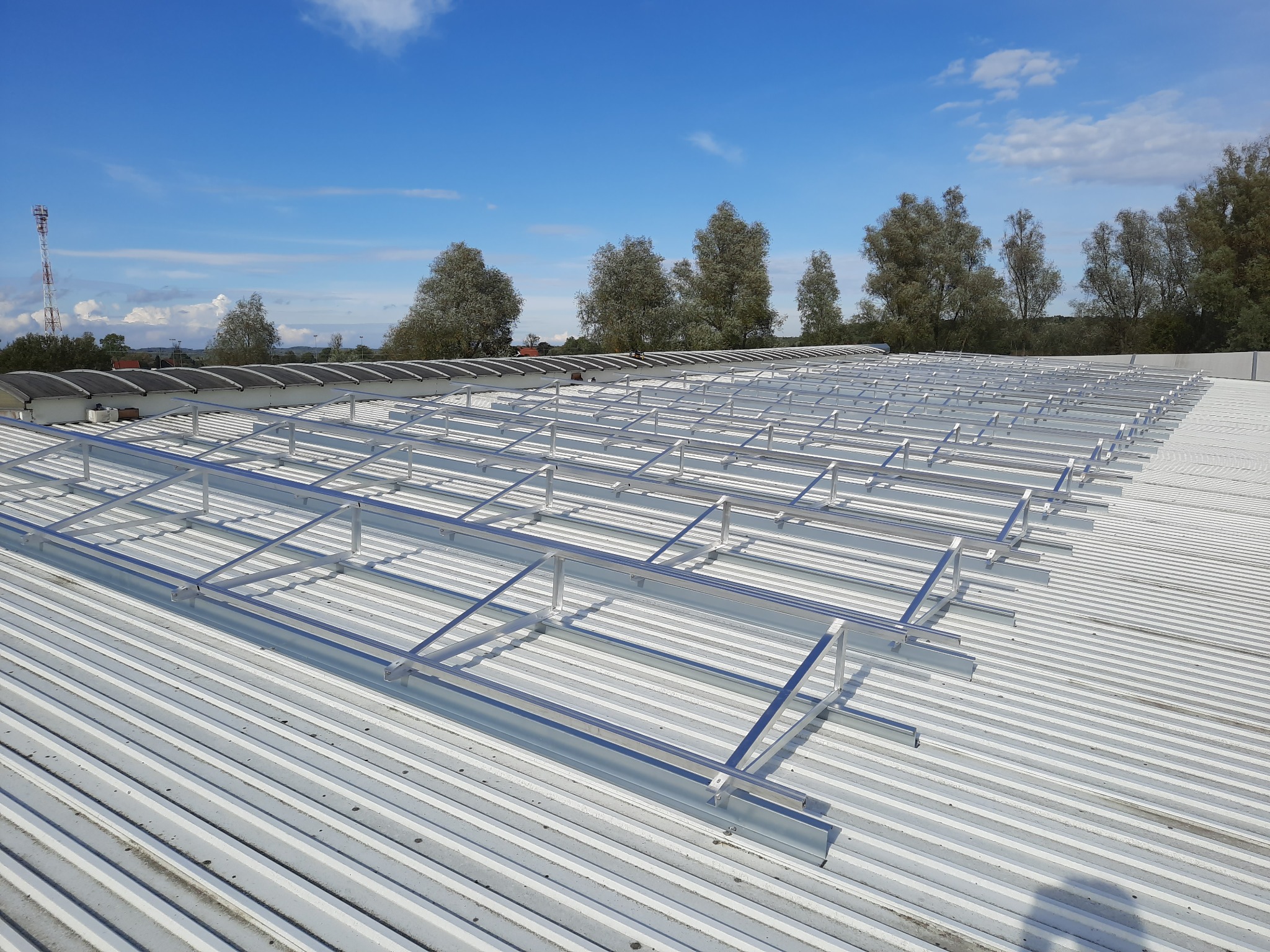 Montaż stelażu pod panele fotowoltaiczne na dachu przemysłowym z blachy trapezowej, widoczne drzewa i wieża telekomunikacyjna w tle.