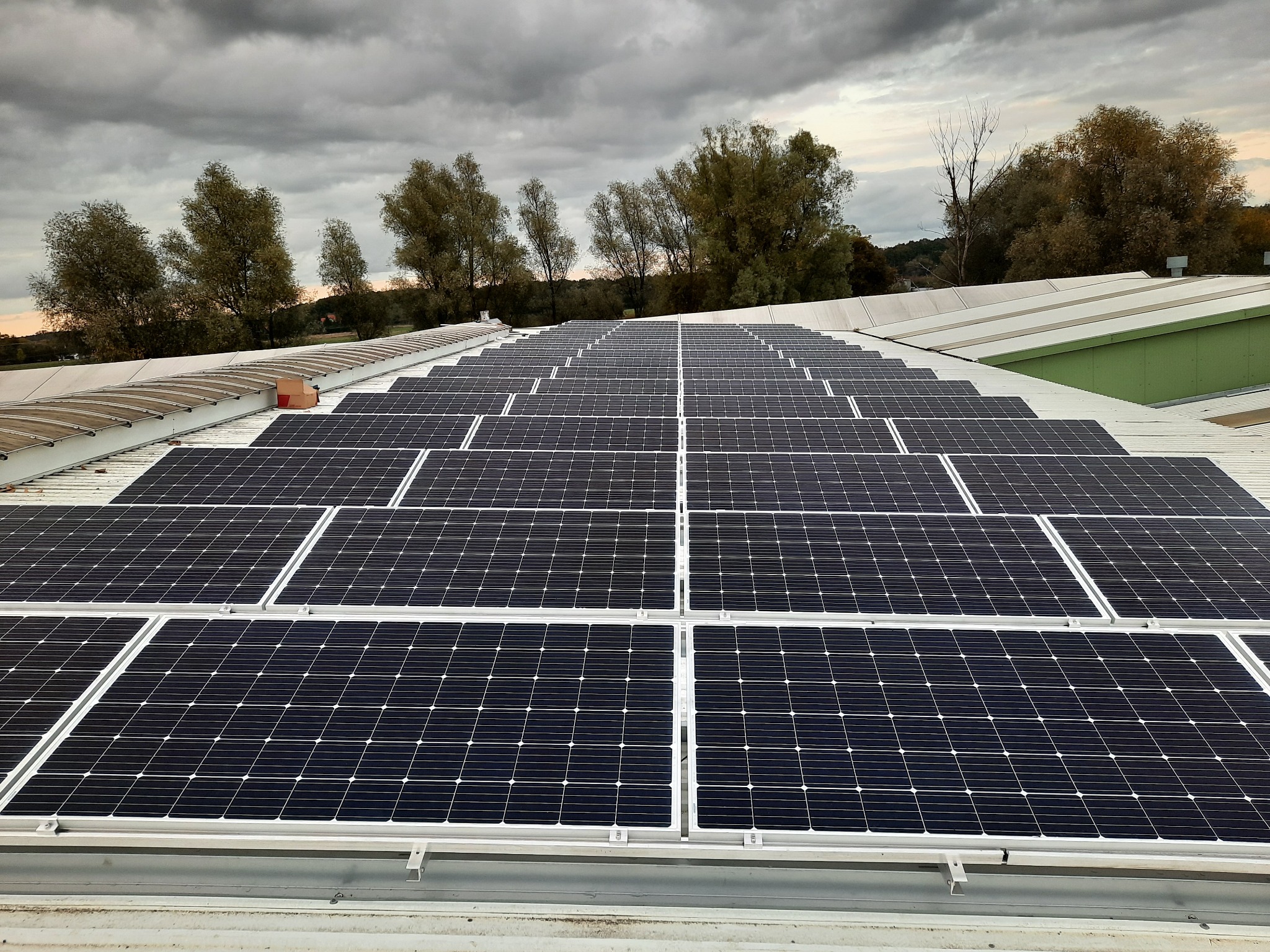 Rząd paneli słonecznych na płaskim dachu budynku przemysłowego, widok z góry przy pochmurnym niebie i zalesionym horyzoncie.
