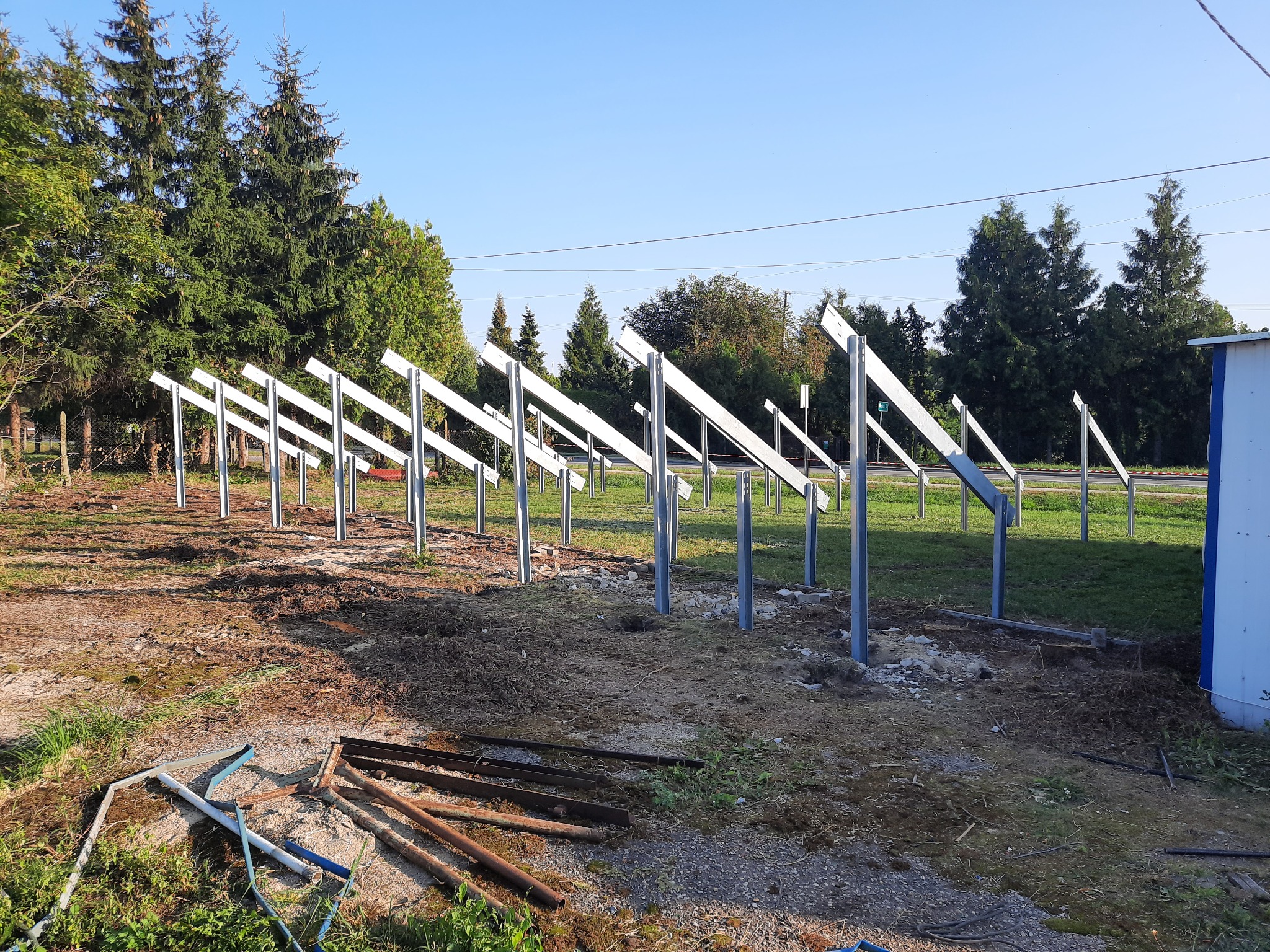 Stalowe konstrukcje wsporcze pod panele słoneczne, ustawione rzędami na przygotowanym terenie, widok z perspektywy ziemi, w tle drzewa i błękitne niebo.