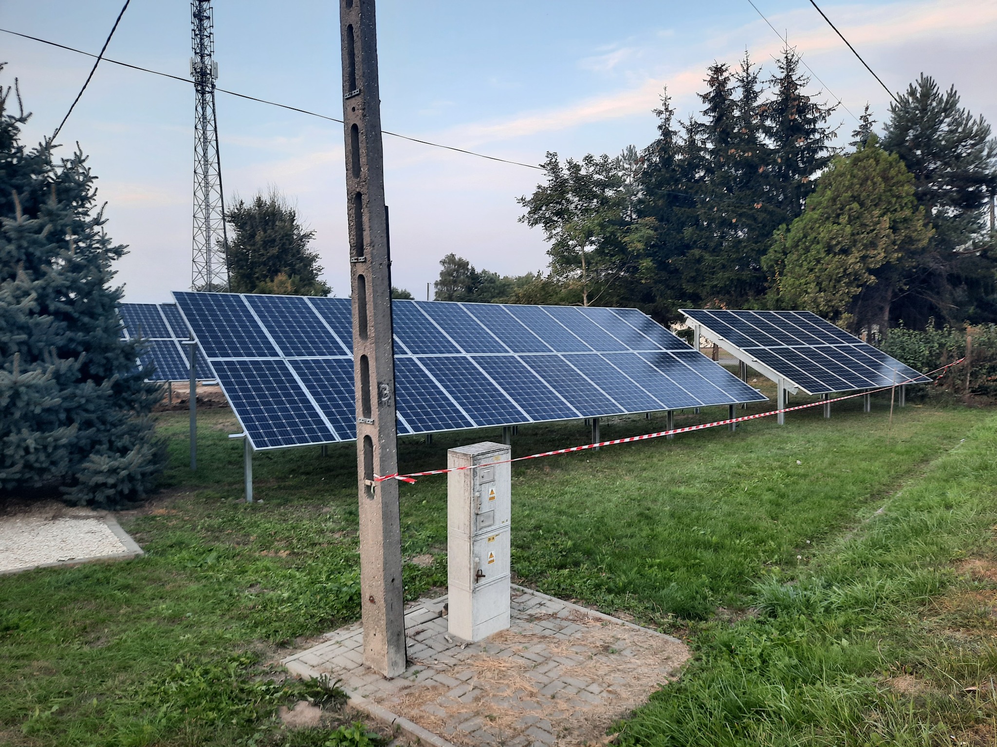 Rząd paneli słonecznych na trawiastym terenie, z widoczną skrzynką elektryczną owiniętą taśmą ostrzegawczą oraz słupem energetycznym w pierwszym planie, w tle drzewa i maszt telekomunikacyjny.