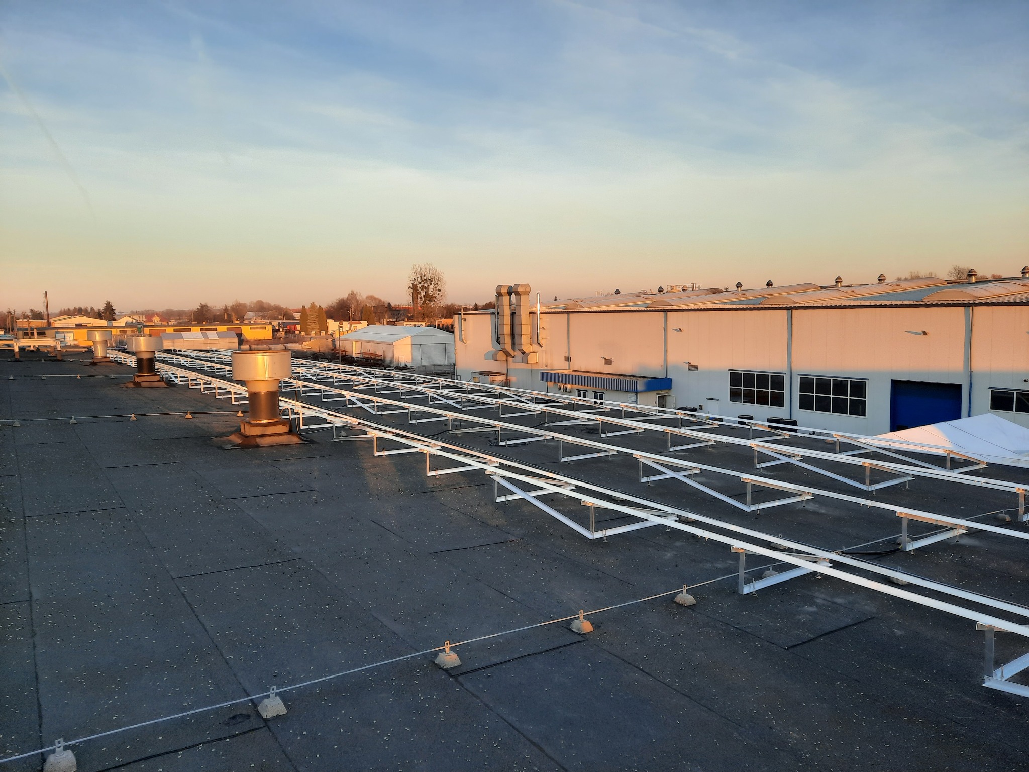 Stelaż pod panele słoneczne na dachu przemysłowym, widoczne elementy montażowe i zabezpieczenia, w tle budynek hali i zachodzące słońce.