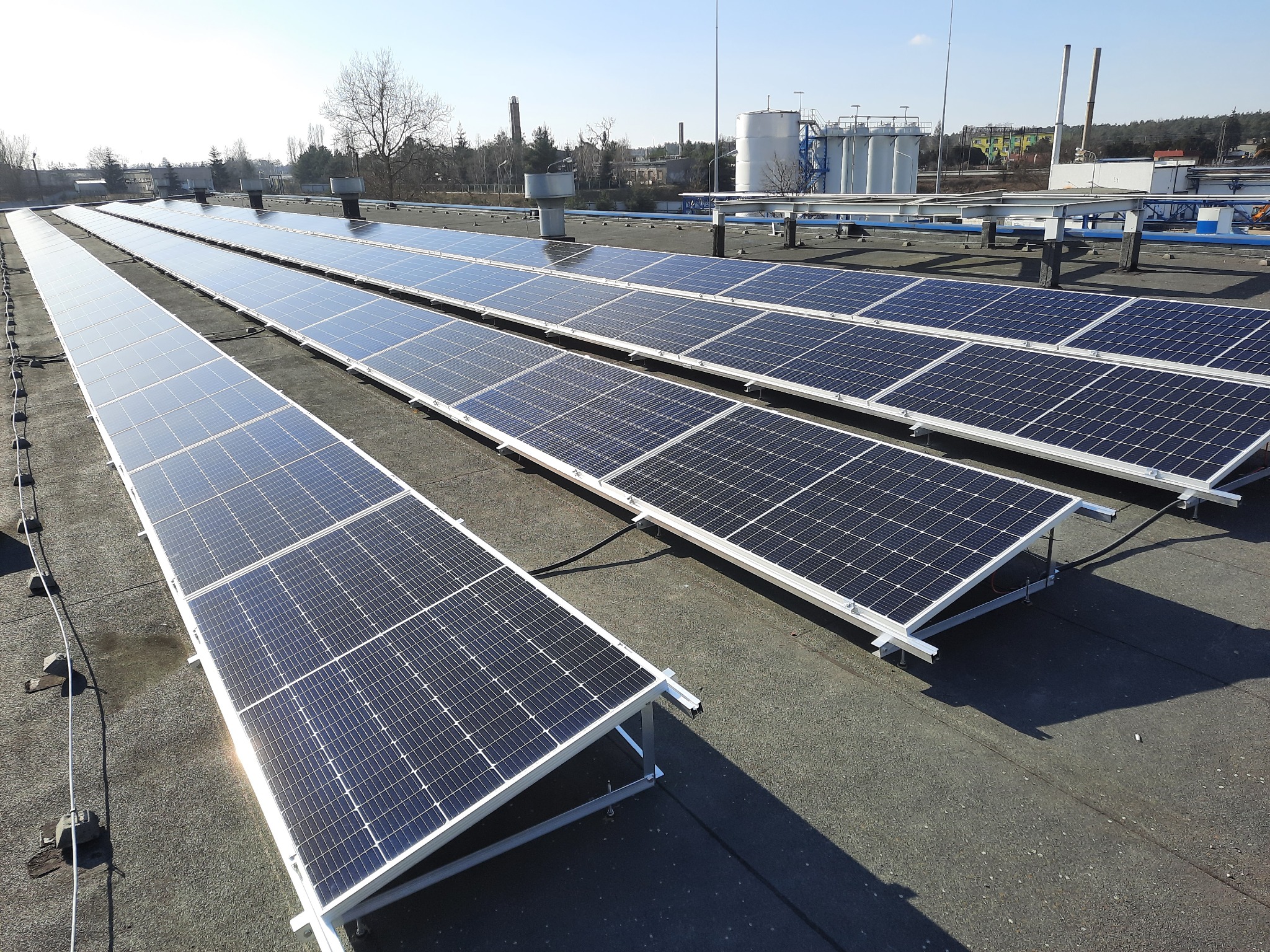 Rząd paneli słonecznych na płaskim dachu budynku przemysłowego, w tle zbiorniki i kominy
