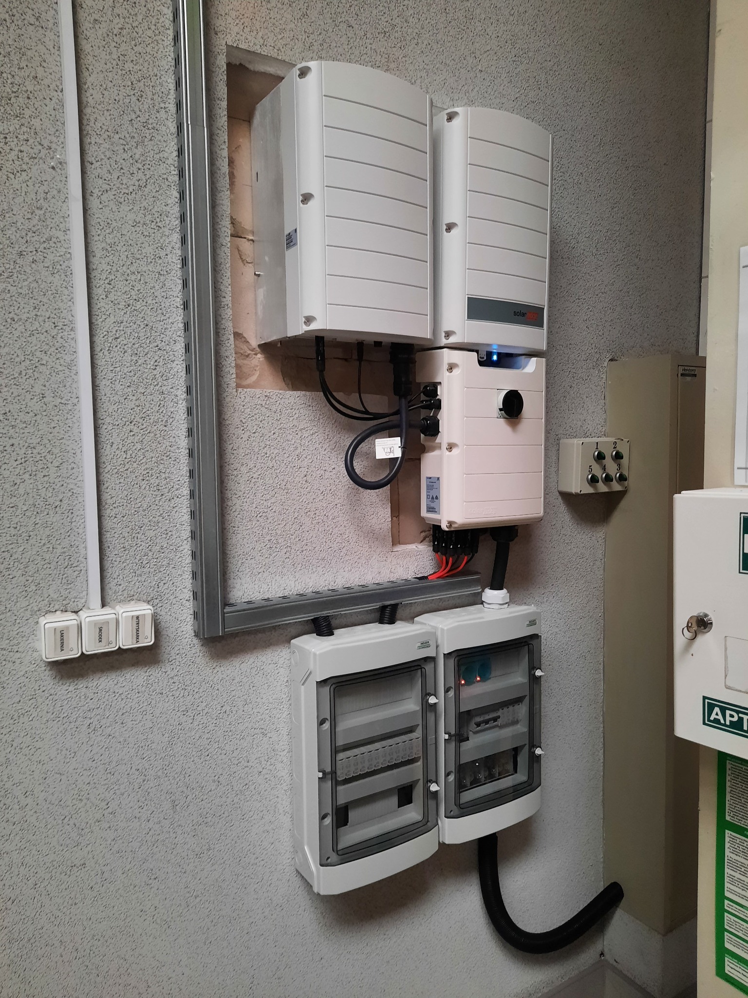 Zamontowane na szorstkiej ścianie, białe falowniki i skrzynki rozdzielcze z widocznymi przewodami, część obudowy skrzynki rozdzielczej jest otwarta.