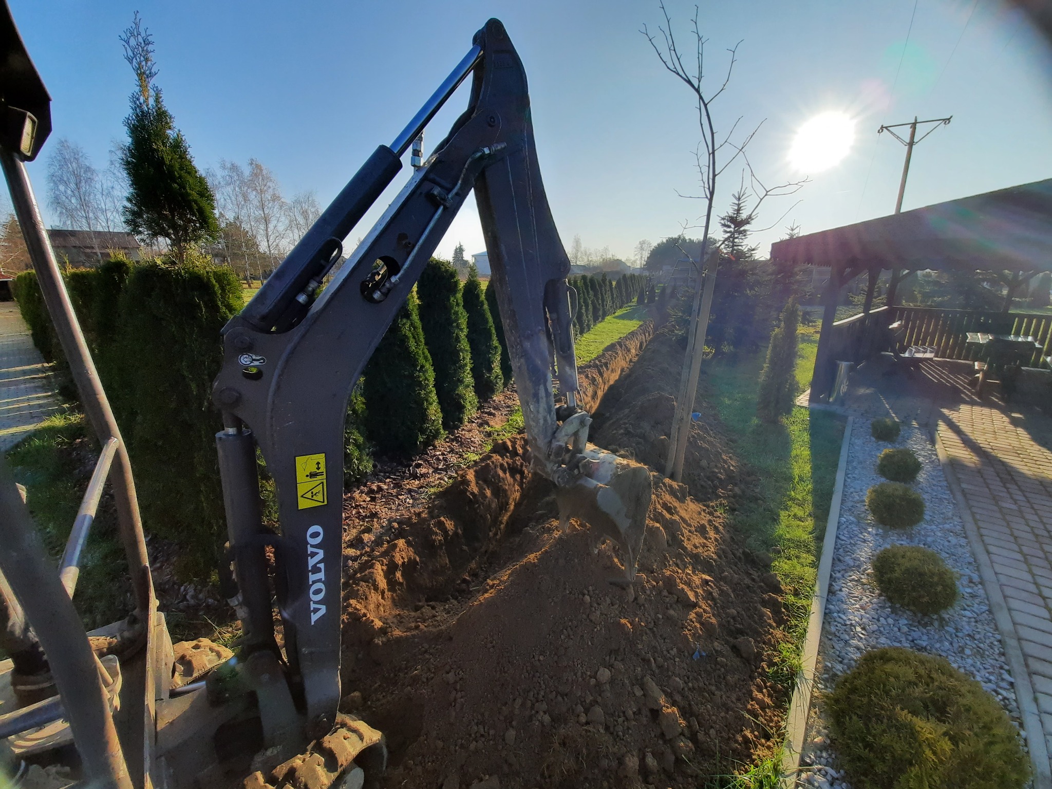 Mini koparka Volvo w trakcie wykopywania wąskiego rowu w ziemi, prawdopodobnie pod instalację, z równo posadzonymi tujami w tle i widocznym zarysem altany.