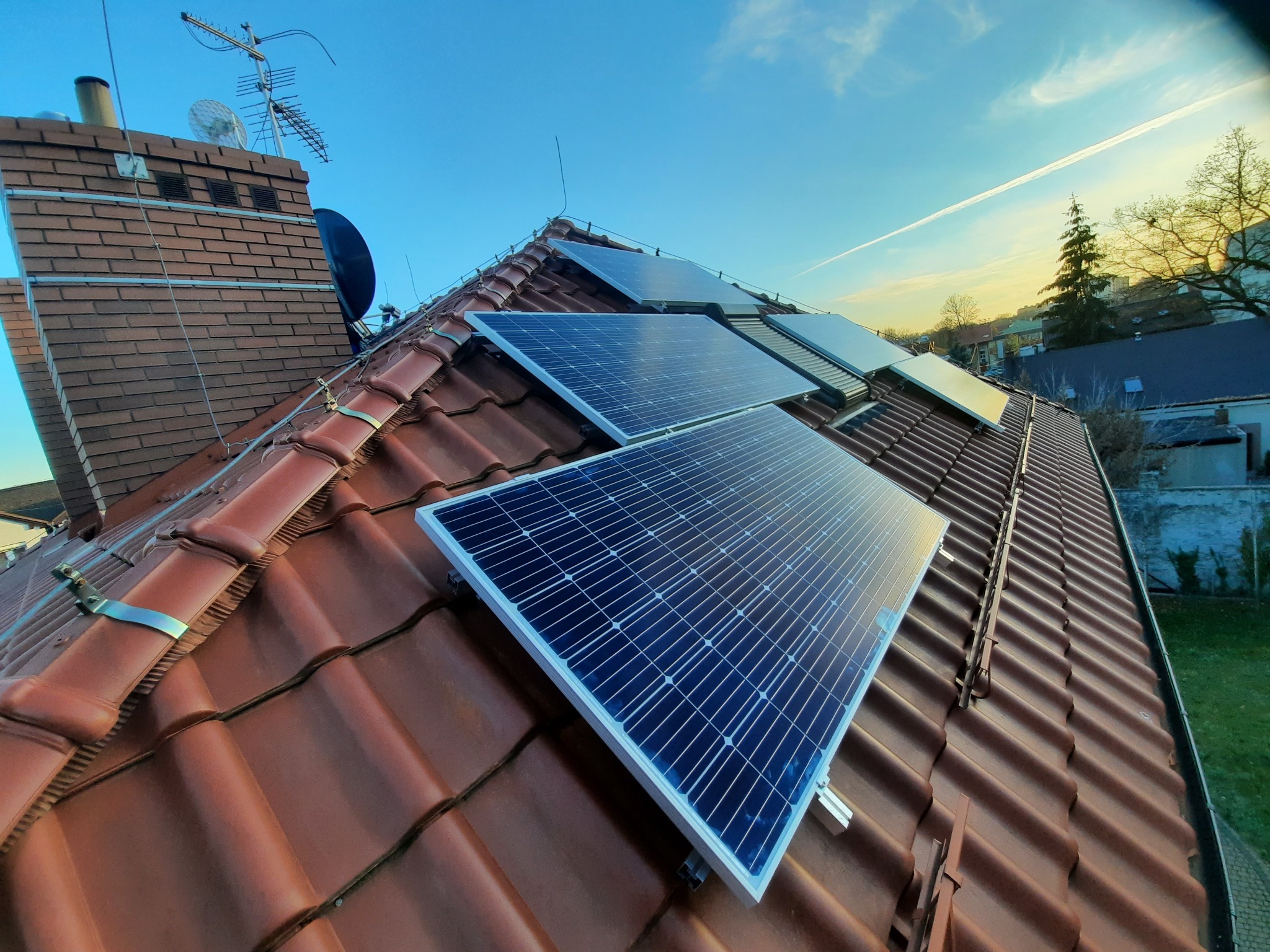 Panele słoneczne zamontowane na czerwonej dachówce, widok z dołu, widoczne mocowania paneli, komin z antenami i błękitne niebo z delikatnymi chmurami w tle.