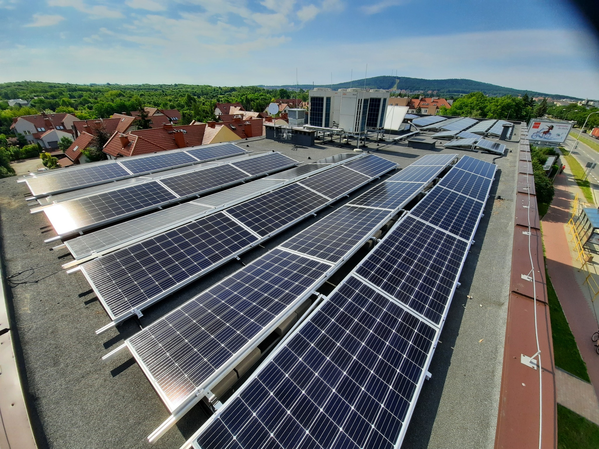 Rząd paneli fotowoltaicznych na płaskim dachu budynku, widok z góry, w tle drzewa, zabudowania i błękitne niebo.