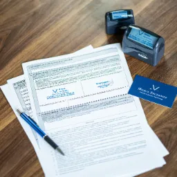 Dokumenty podatkowe, pieczątki firmowe i wizytówka Kancelarii Doradcy Podatkowego Michała Zbutowicza leżące na drewnianym biurku, niebieski długopis obok.