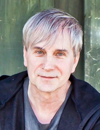 Portret mężczyzny o jasnych włosach i niebieskich oczach, ubrany w ciemną kurtkę na tle zielonych drzwi.