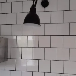 Czarna, metalowa lampa ścienna w stylu industrialnym zamontowana na białej ścianie z płytek typu metro z czarną fugą, widoczne gniazdko elektryczne i fragment blatu.